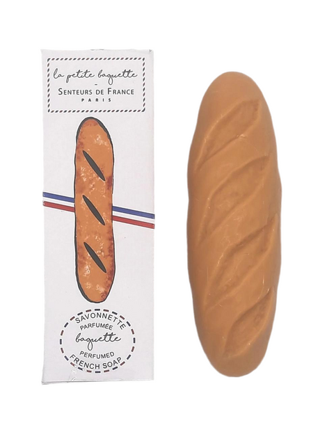 A baguette-shaped soap with a beige color, placed next to its packaging which has 'la petite baguette' written on it, against a white background.