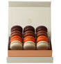 A boxed assortment of 12 French macarons arranged in three columns inside an open cream-colored gift box with a rose-gold interior base. The macarons include dark chocolate, milk chocolate, caramel brown, orange, and vanilla flavors.