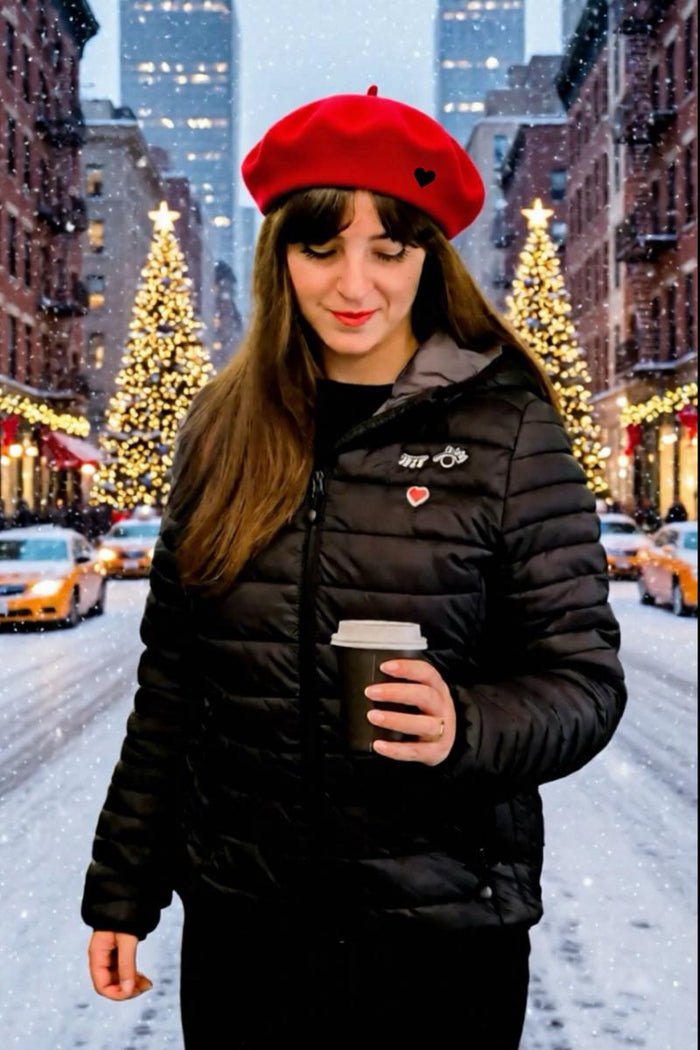 French Woman in a red beret and black puffer jacket holding a coffee cup on a snowy New York city street with Christmas lights.