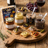 Assorted jars of condiments on a wooden board with bread slices and pickles, set on a rustic wooden table.