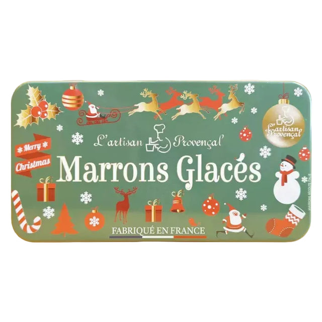 A rectangular metal tin of L'Artisan Provençal Marrons Glacés, decorated with a festive Christmas theme on a muted green background. The tin features illustrations of Santa Claus, reindeer, a snowman, gifts, and candy canes, with the brand name 'L'Artisan Provençal' and product name 'Marrons Glacés' printed in white.