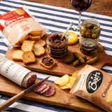 aperitif Platter with chips, spread, pickles, and charcuterie on a striped tablecloth.