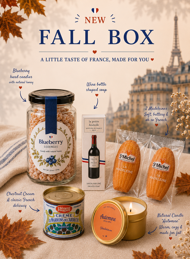 A styled fall gift box arrangement on a beige fabric surface with autumn leaves and a blurred Eiffel Tower background. Visible items include a large jar labeled Blueberry Hard Candles, a wine-shaped soap box, two St.