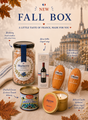 A styled fall gift box arrangement on a beige fabric surface with autumn leaves and a blurred Eiffel Tower background. Visible items include a large jar labeled Blueberry Hard Candles, a wine-shaped soap box, two St.