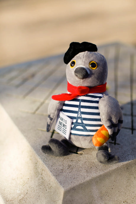 Stuffed toy pigeon in Paris-themed outfit with a red scarf and black beret, holding an orange, on a concrete surface.