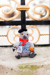 Plush toy pigeon in a Parisian outfit with a red scarf and striped shirt, standing in front of an ornate metal gate.