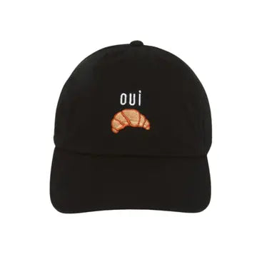 A black baseball cap featuring an embroidered white cursive word 'oui' and a small orange croissant graphic on the front, set against a plain white background.