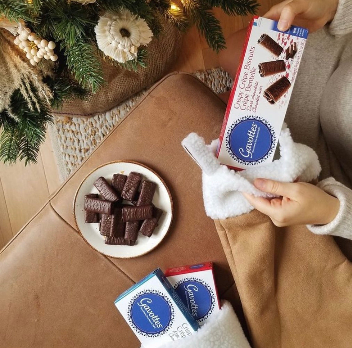 Overhead lifestyle shot of Gavottes dark chocolate crispy crêpes served on a white ceramic plate beside Christmas tree décor, with a person holding the red dark chocolate box and the blue milk chocolate box on the floor